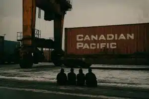 The Protest Is Over. Now What? | John Prince A small group of silhouetted people sit in front of a massive container crane at a port or rail yard, blocking the machinery.