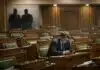 Parliament in Name Only: Representation Without Power Parliament in name only. A single MP sitting alone in a nearly empty House of Commons, head down, looking at a phone, with party whips visible in shadow in the background.