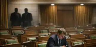 Parliament in name only. A single MP sitting alone in a nearly empty House of Commons, head down, looking at a phone, with party whips visible in shadow in the background.